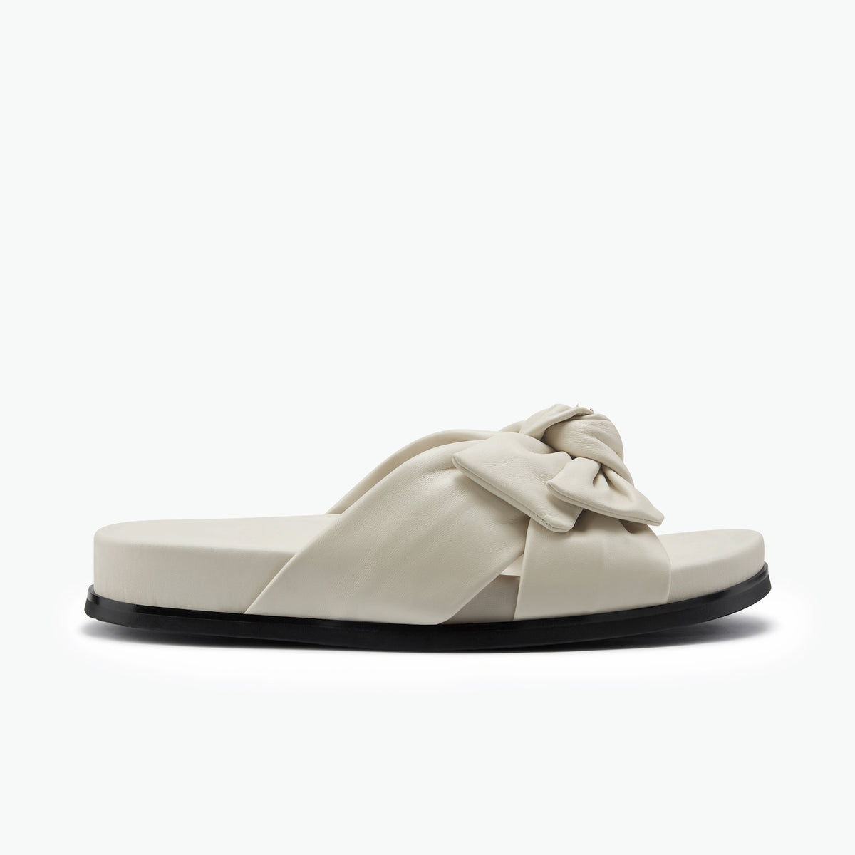A single flat slide sandal with a wide, padded crisscross strap featuring a large bow detail, shown in a light cream color with a black sole on a white background.
