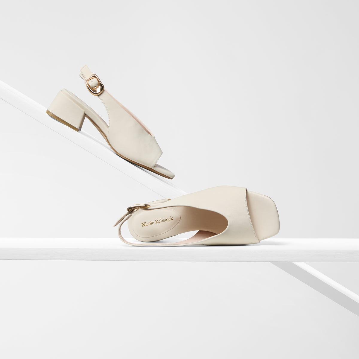 A pair of ivory leather peep-toe block-heel sandals with ankle straps and gold-tone buckles, displayed on a minimalist white geometric set. One shoe is shown from a top-down angle on a diagonal platform, and the other is positioned below on a horizontal surface, with the brand name 'Nicole Rebstock' visible on the insole.