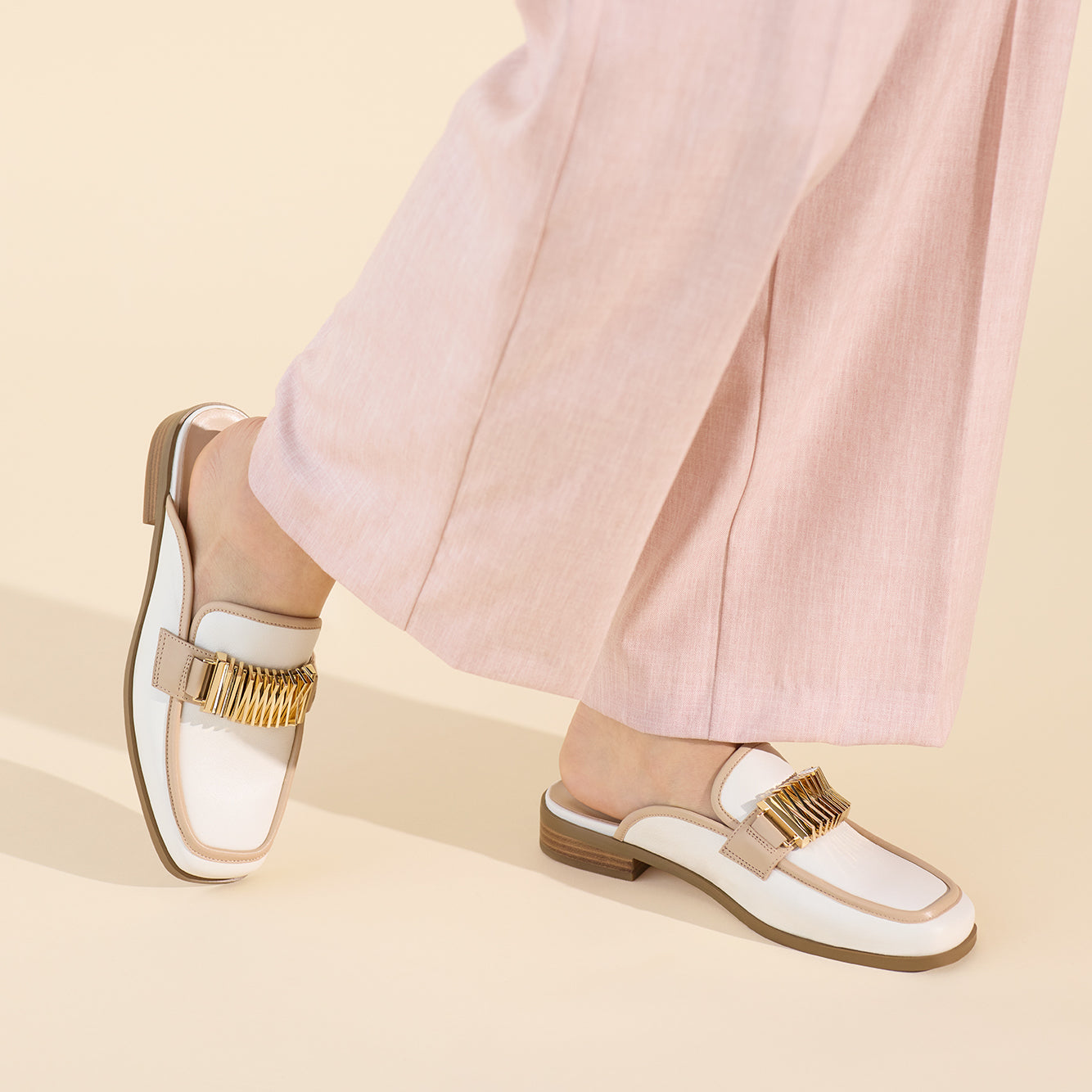White loafers with nude and gold accents worn by a person in light pink pants on a beige background