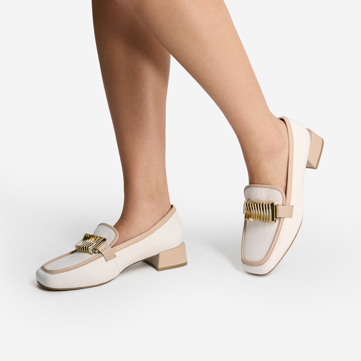 White loafers with nude and gold accents worn by a person on a light grey background. Square toe.