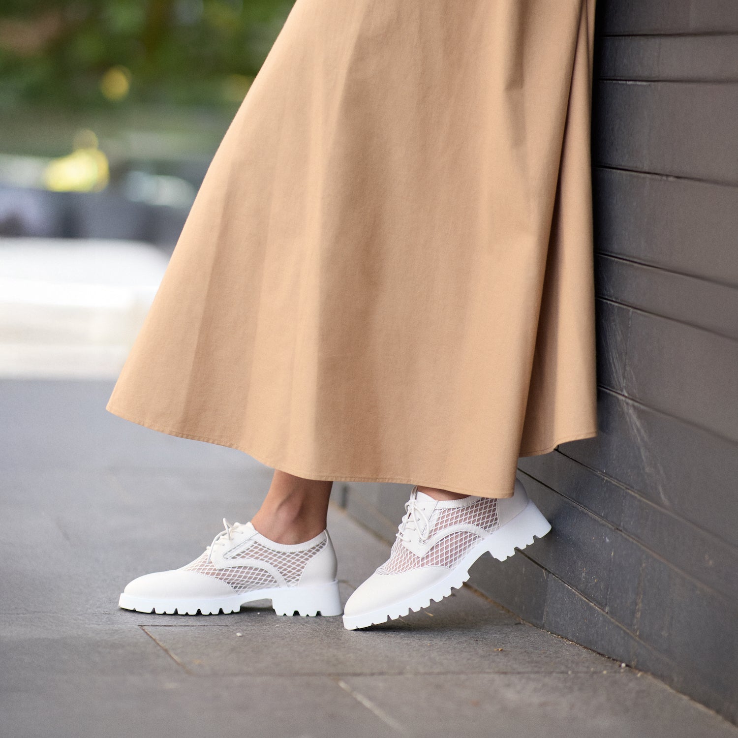 Person wearing a beige coat and white flats leaning against a wall.