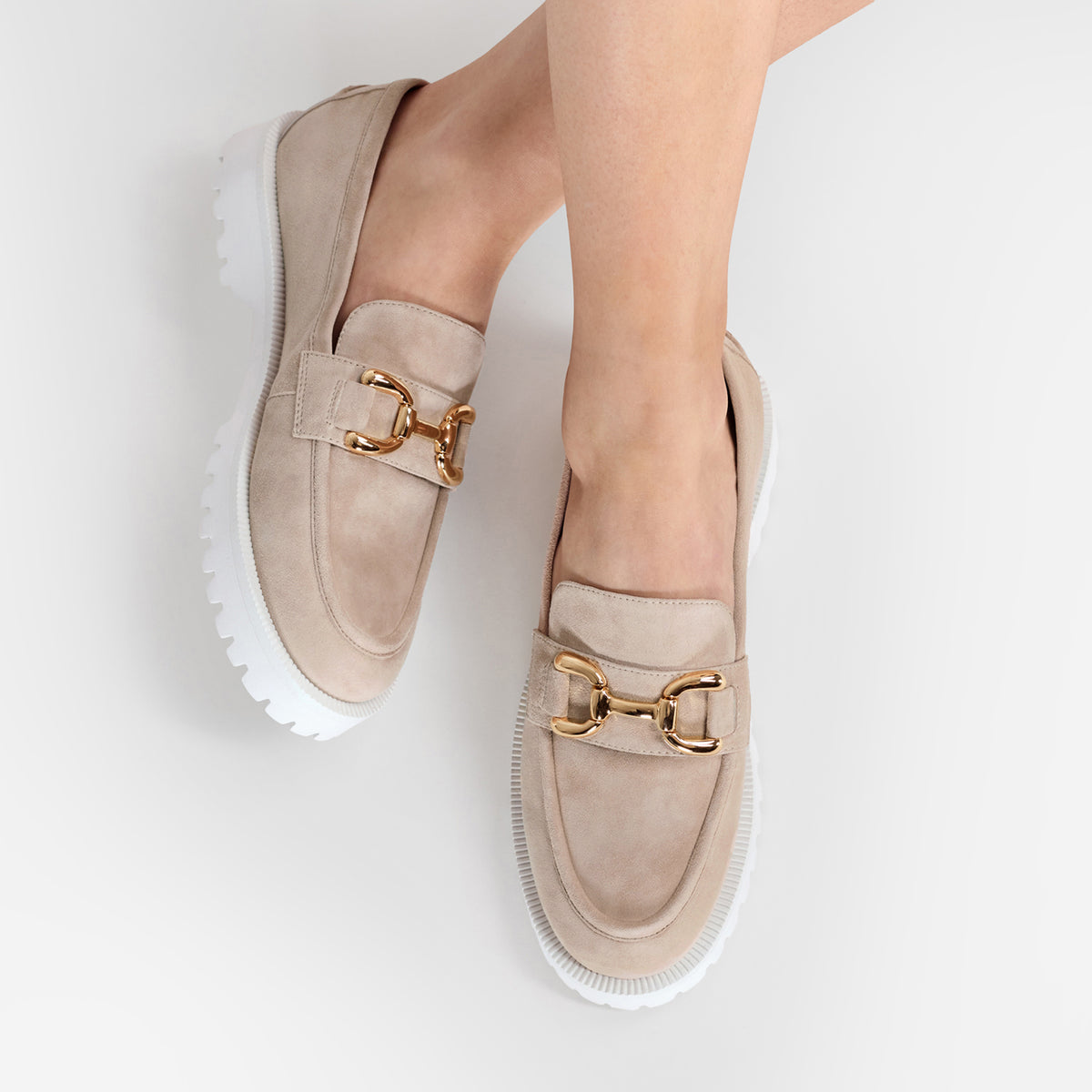 A pair of beige suede loafers with chunky white platform soles and gold-tone horsebit hardware, shown on a model's feet against a light gray background.