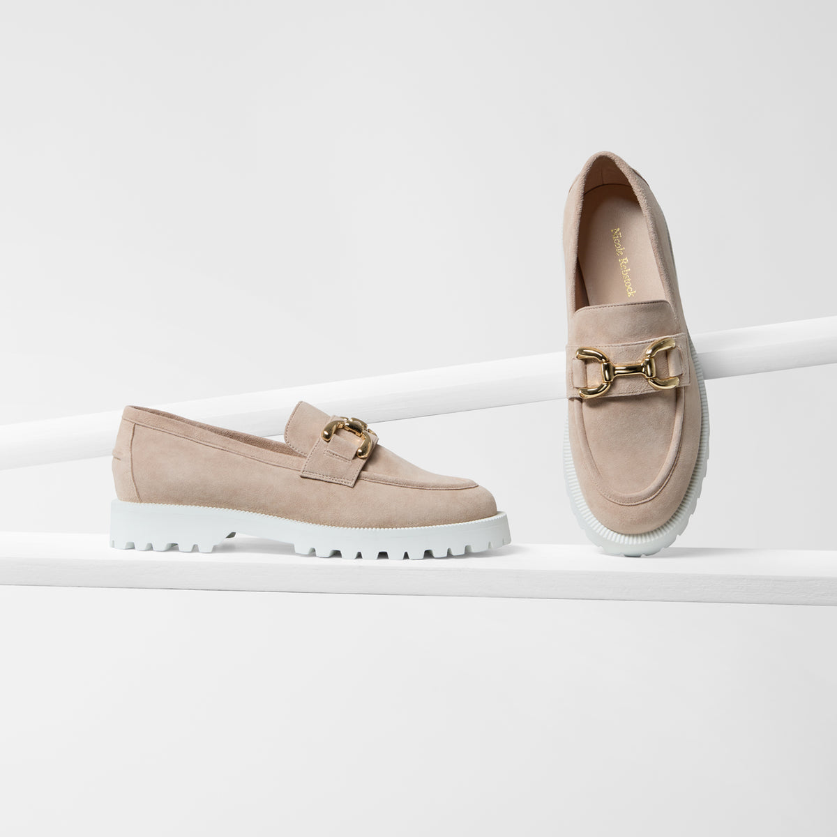 A pair of beige slip-on loafers with chunky white soles and gold-tone horsebit hardware by Nicole Rebstock, displayed on a clean white studio set.