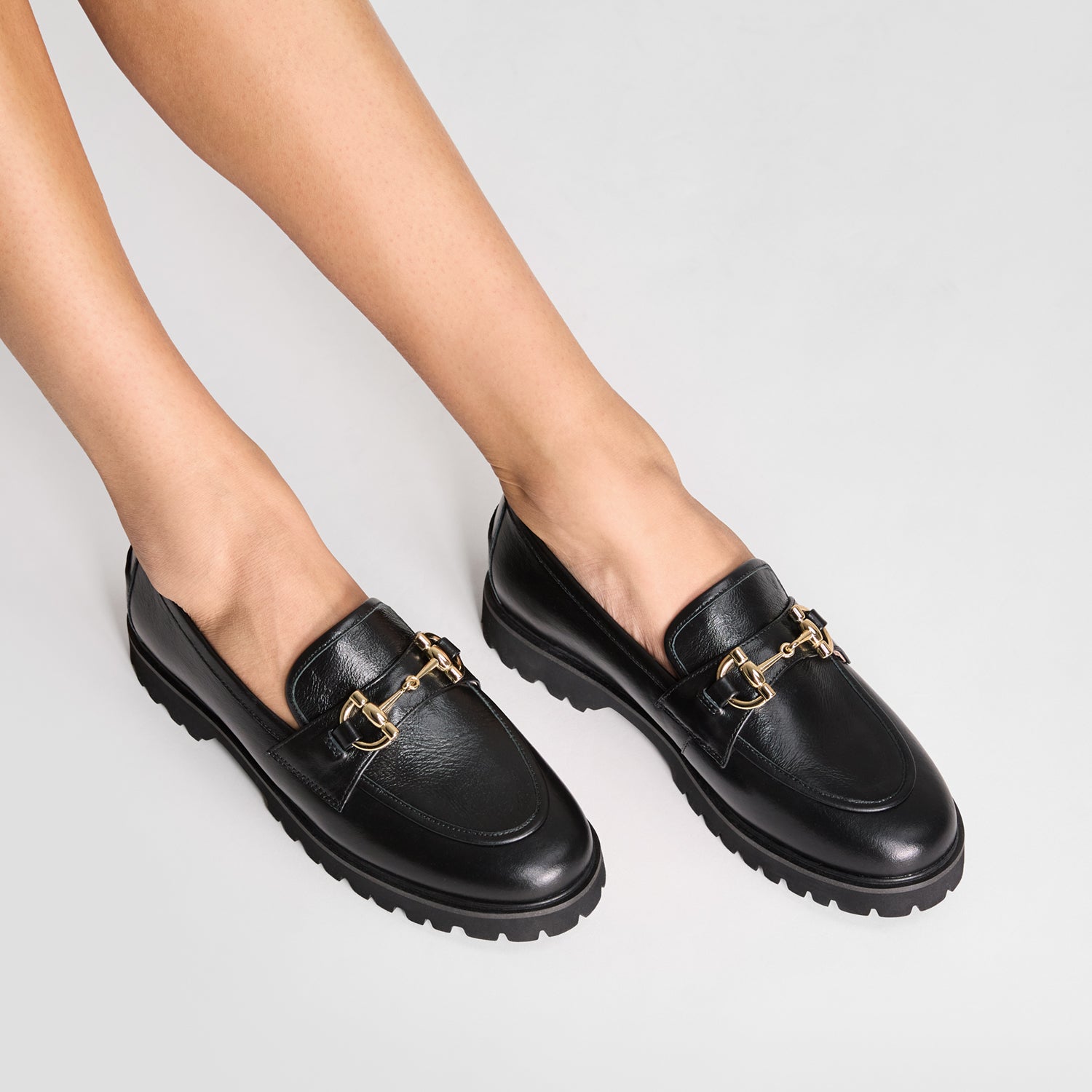 Black loafers with gold accents worn by a person on a light gray background by Nicole Rebstock