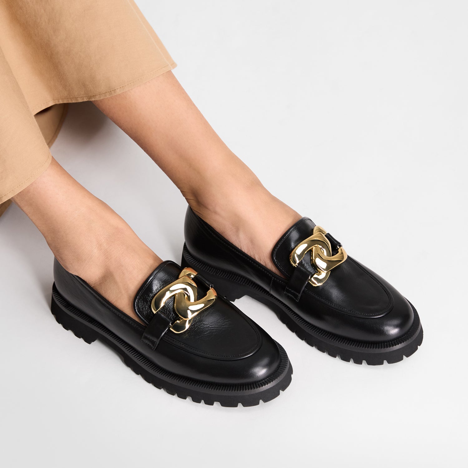 Black loafers with gold chain details worn by a person on a light grey background. Chunky sole. 2cm heel.