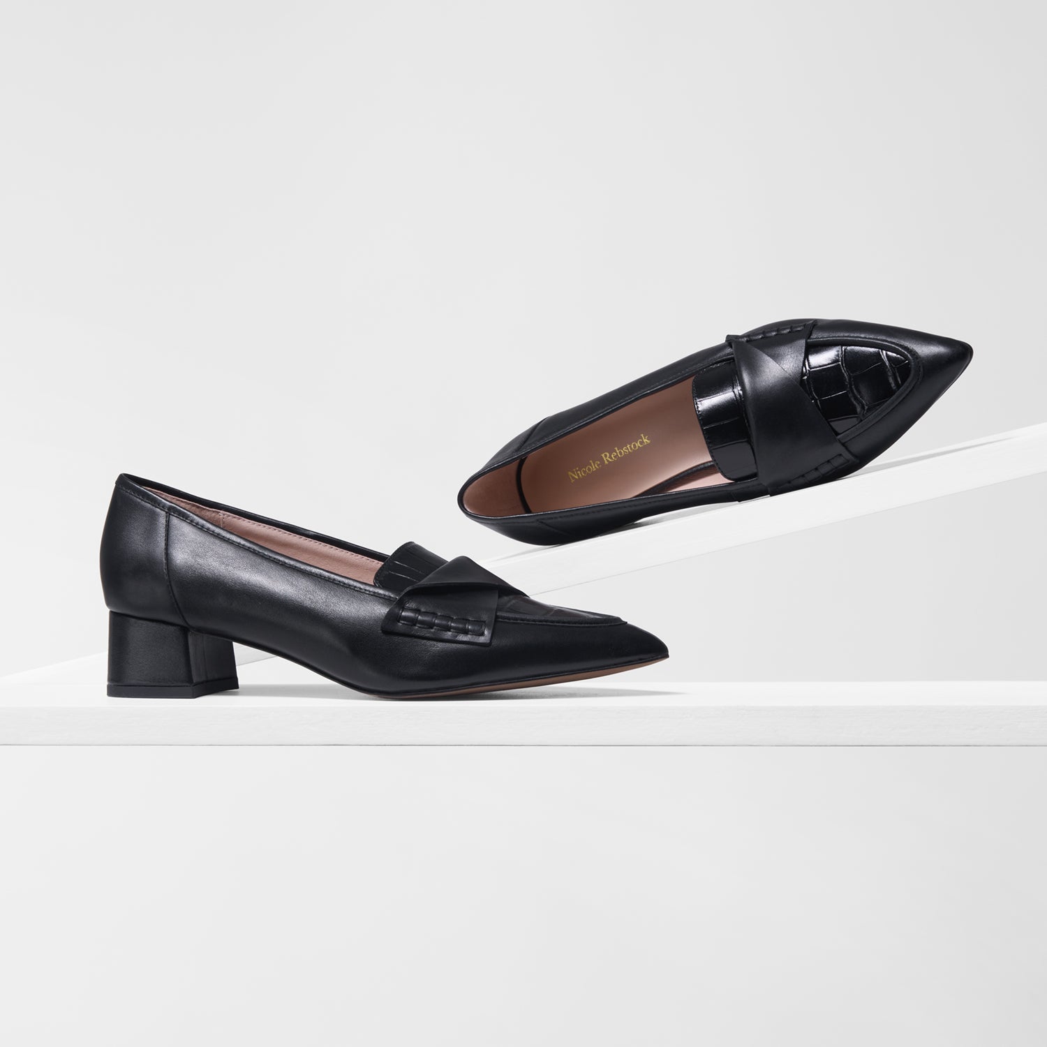 A pair of black leather loafers with low block 4cm heels, shown on a clean white platform against a light gray background. One shoe is positioned flat on the platform, and the other is angled above it to show the pointed toe and decorative side detail.