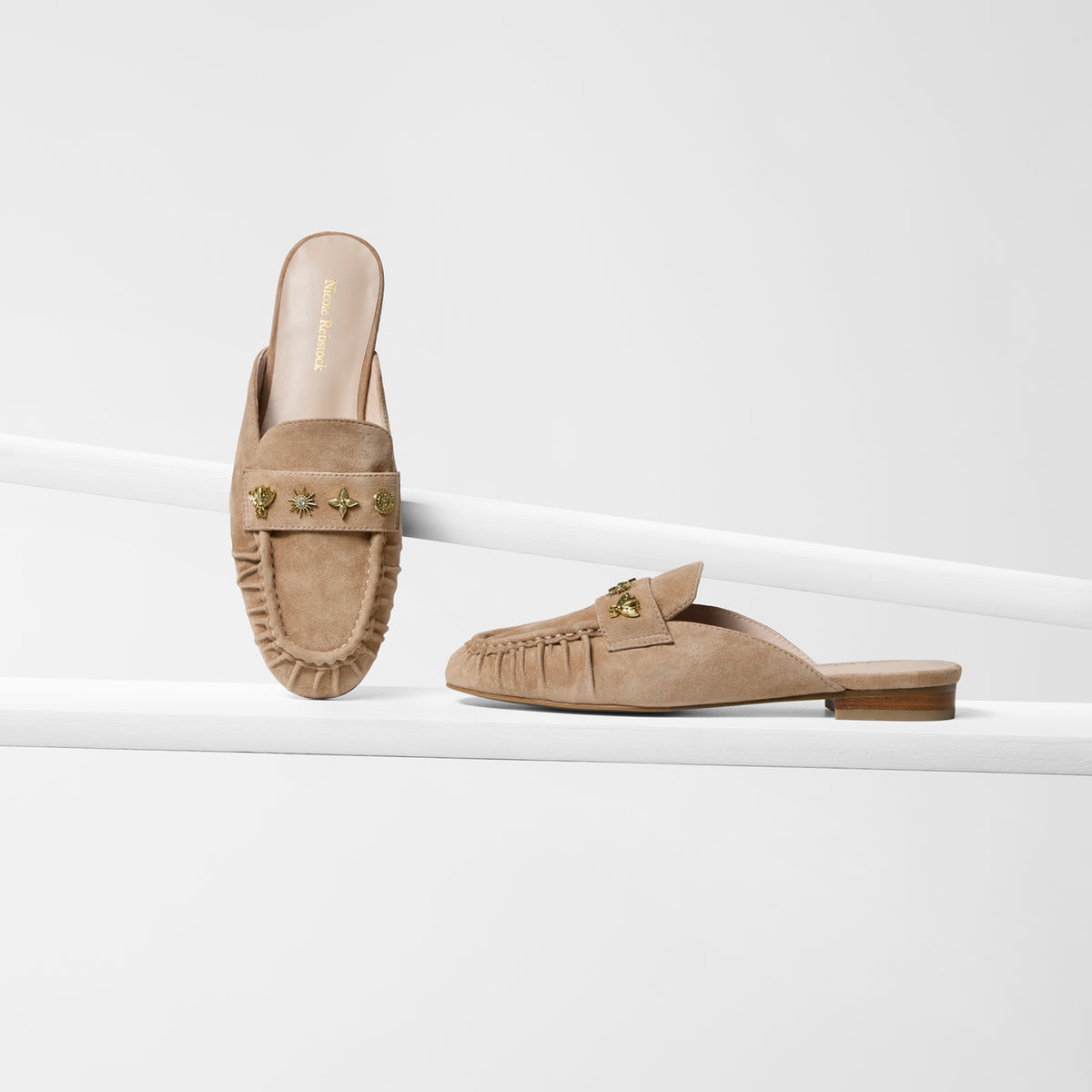 A pair of flat nude suede loafer mules with gold studs by Nicole Rebstock shown on a grey background from various angles.
