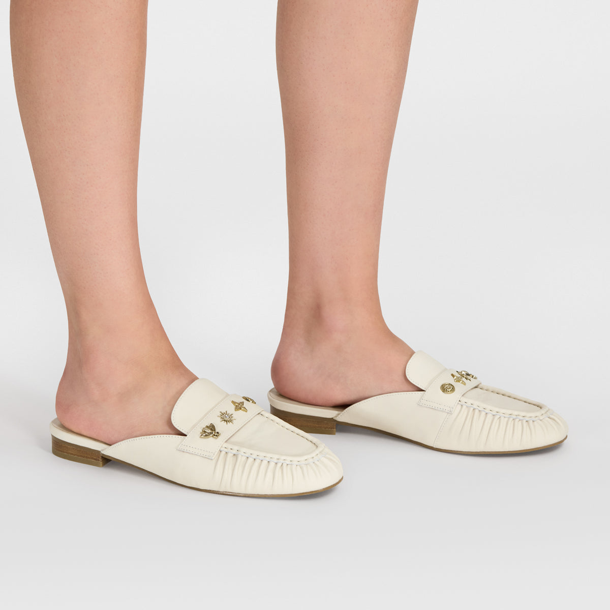 A pair of ivory leather slip-on mule loafers with gold-tone decorative hardware, shown on a model's feet against a light gray studio background.