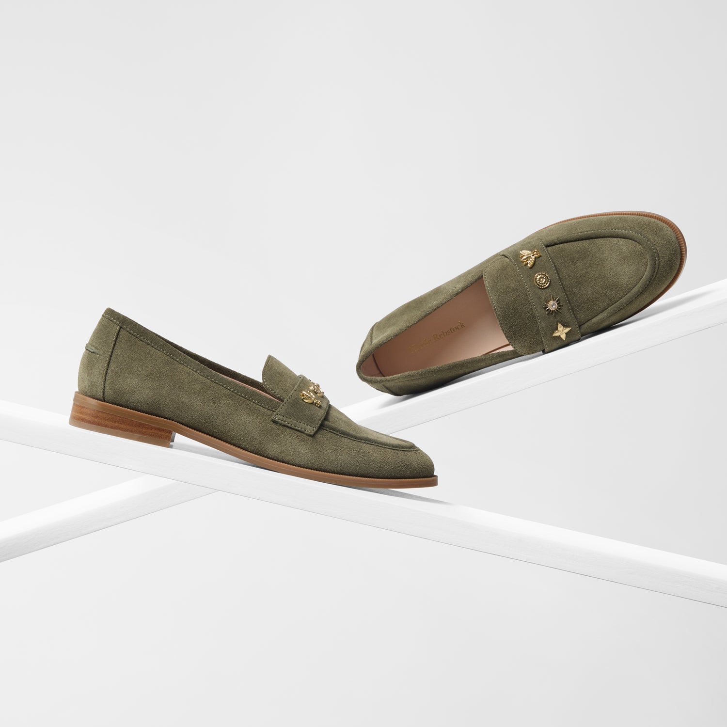 A pair of sage green suede penny loafers with unique gold-tone star stud embellishments on the vamp, displayed on a clean white geometric riser.