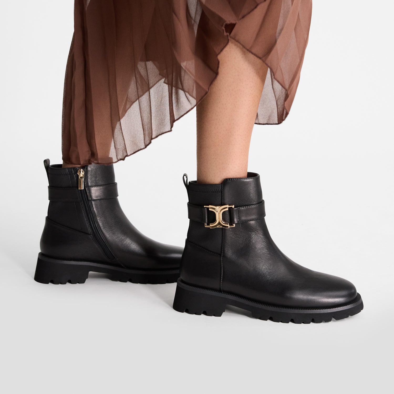 Black flat ankle boots with chunky thread and gold buckle on a white background worn by a model.