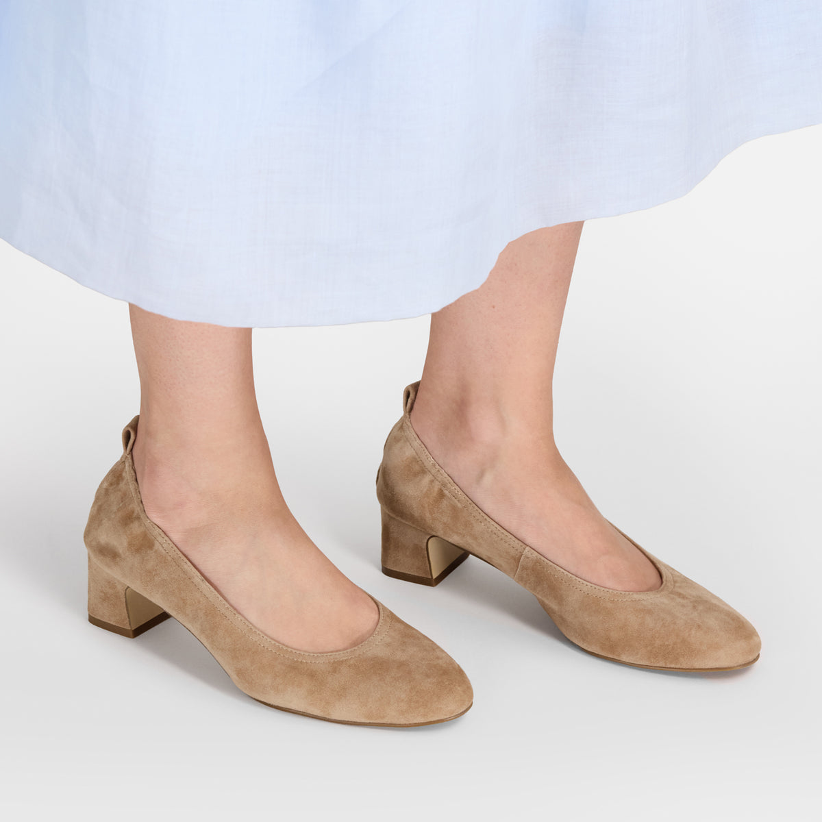 A pair of women's taupe suede block-heel pumps shown on a model's feet, with a light blue skirt hem visible at the top against a clean white studio background.
