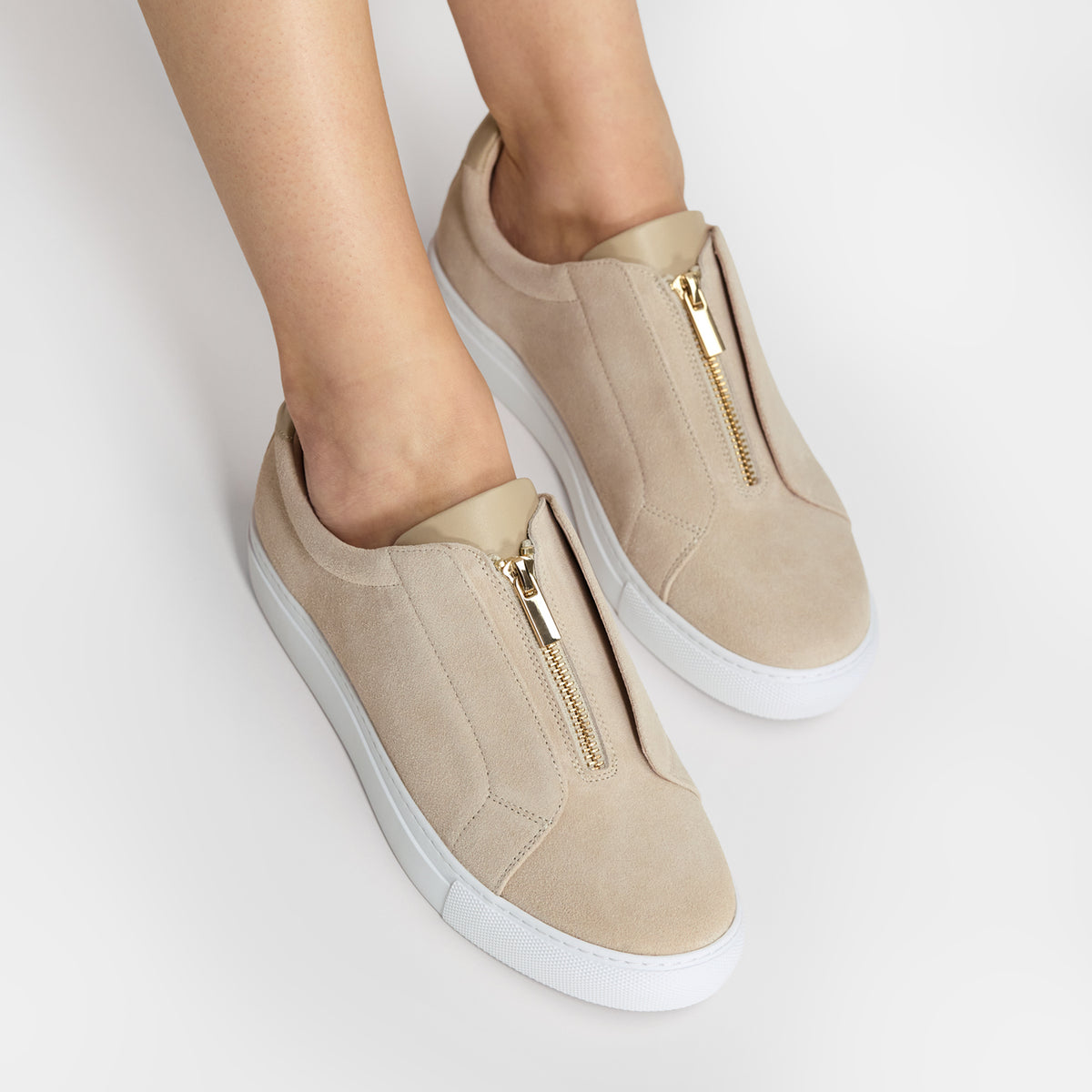 Beige slip-on sneakers with gold-tone front zippers and white soles, shown on a model's feet against a light gray studio background.