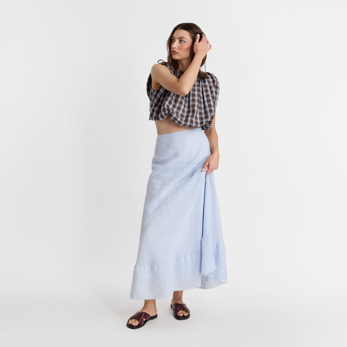 A woman modeling a light blue linen maxi skirt paired with a brown and white checkered crop top and dark brown slide sandals, set against a plain white studio background.