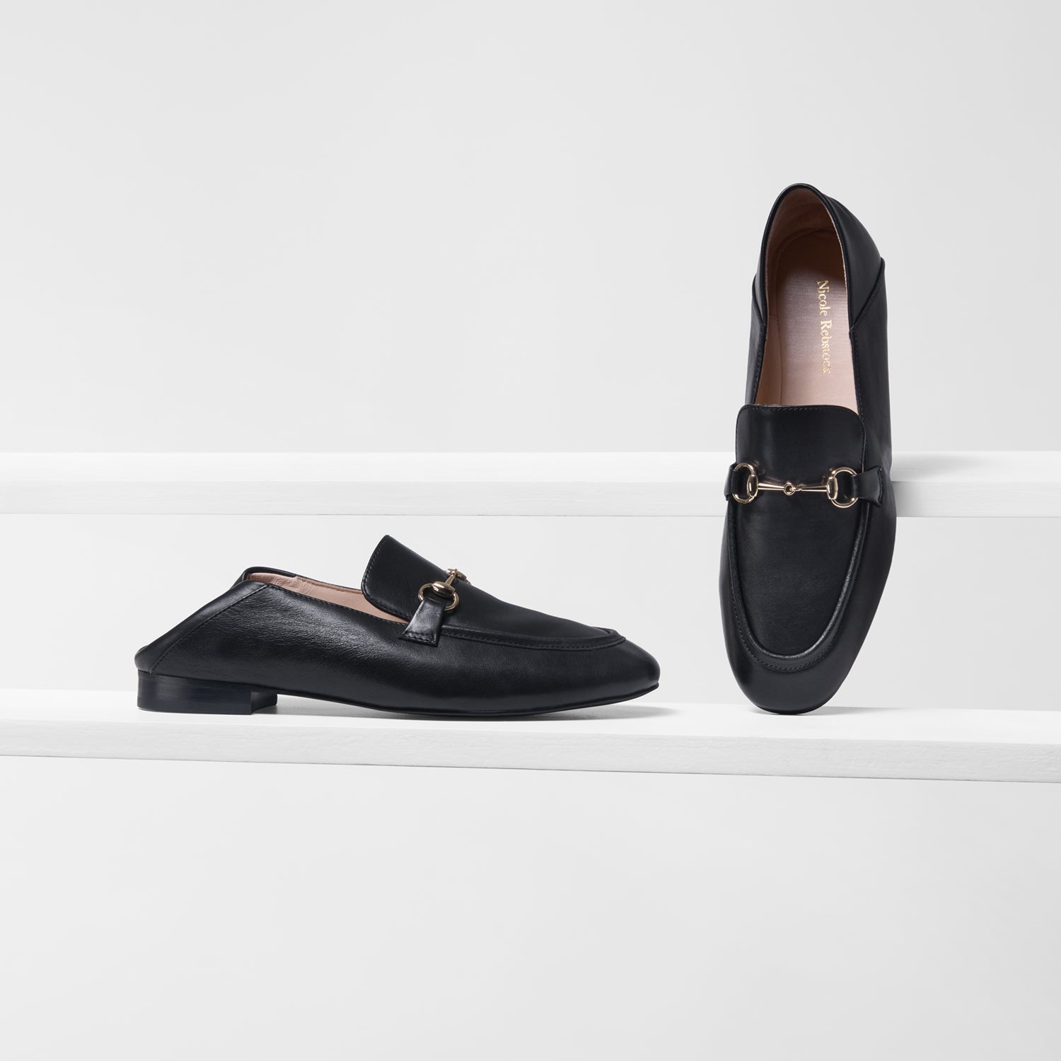 A pair of black leather loafers with gold-tone horsebit detailing, displayed on white steps against a light gray background.