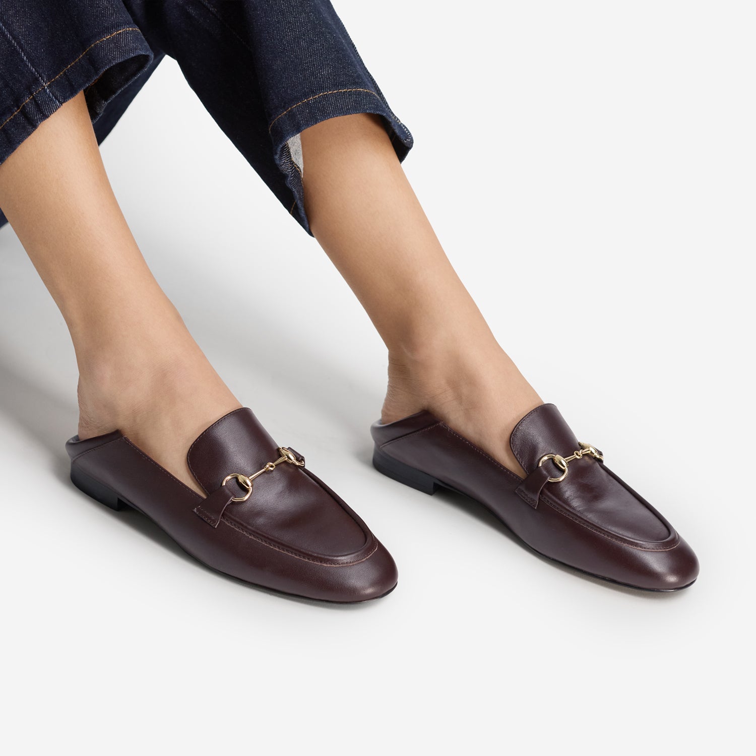 A person wearing dark brown leather slip-on loafers with a gold horsebit detail, shown from the feet up against a light gray studio background.