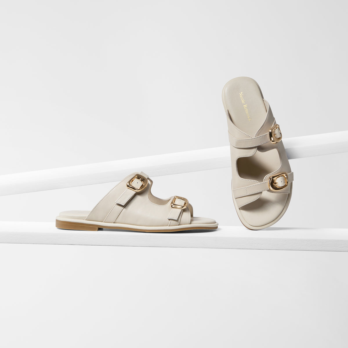 A pair of women's double-strap flat sandals in light beige leather with gold-tone buckle details, displayed on a clean white geometric riser.