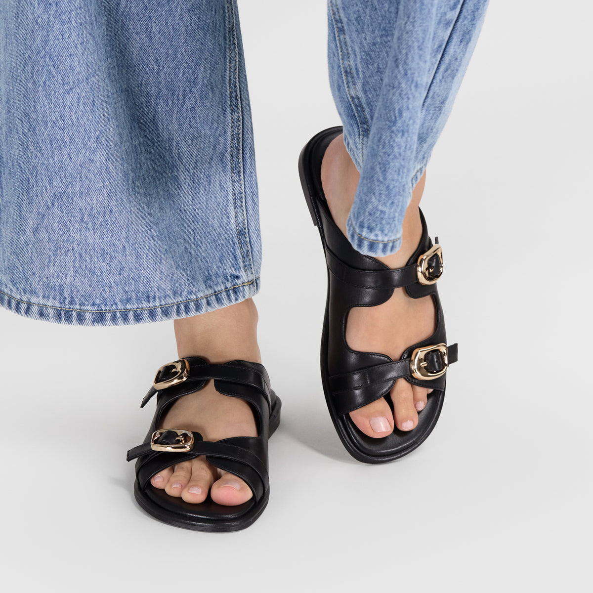 A person wearing black double-strap leather sandals with gold-tone buckle details, paired with light-wash blue jeans, shown from the calves down on a light gray studio background.