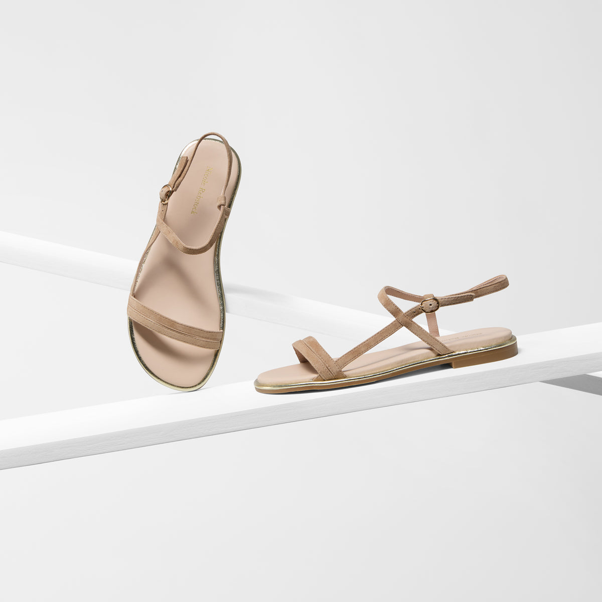A pair of minimalist flat sandals in a light tan or beige suede leather material, displayed on a clean white geometric riser. One sandal is shown upright and the other is laid flat to display the strap configuration.