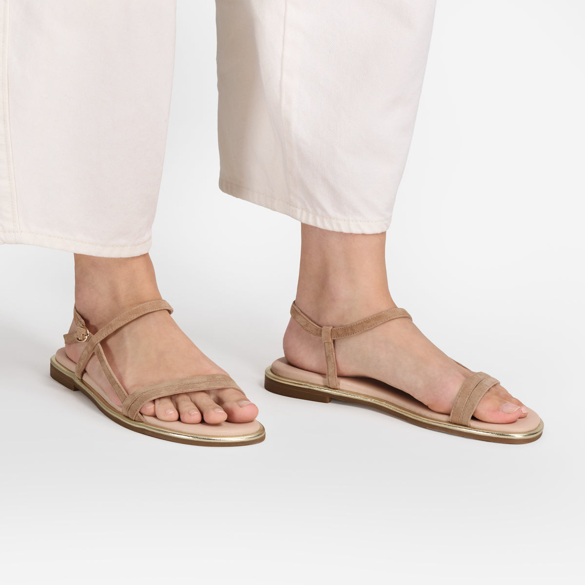 A person wearing beige flat strappy sandals with a metallic gold-toned sole edge, paired with off-white cropped pants, shown from the calves down on a clean white studio background.