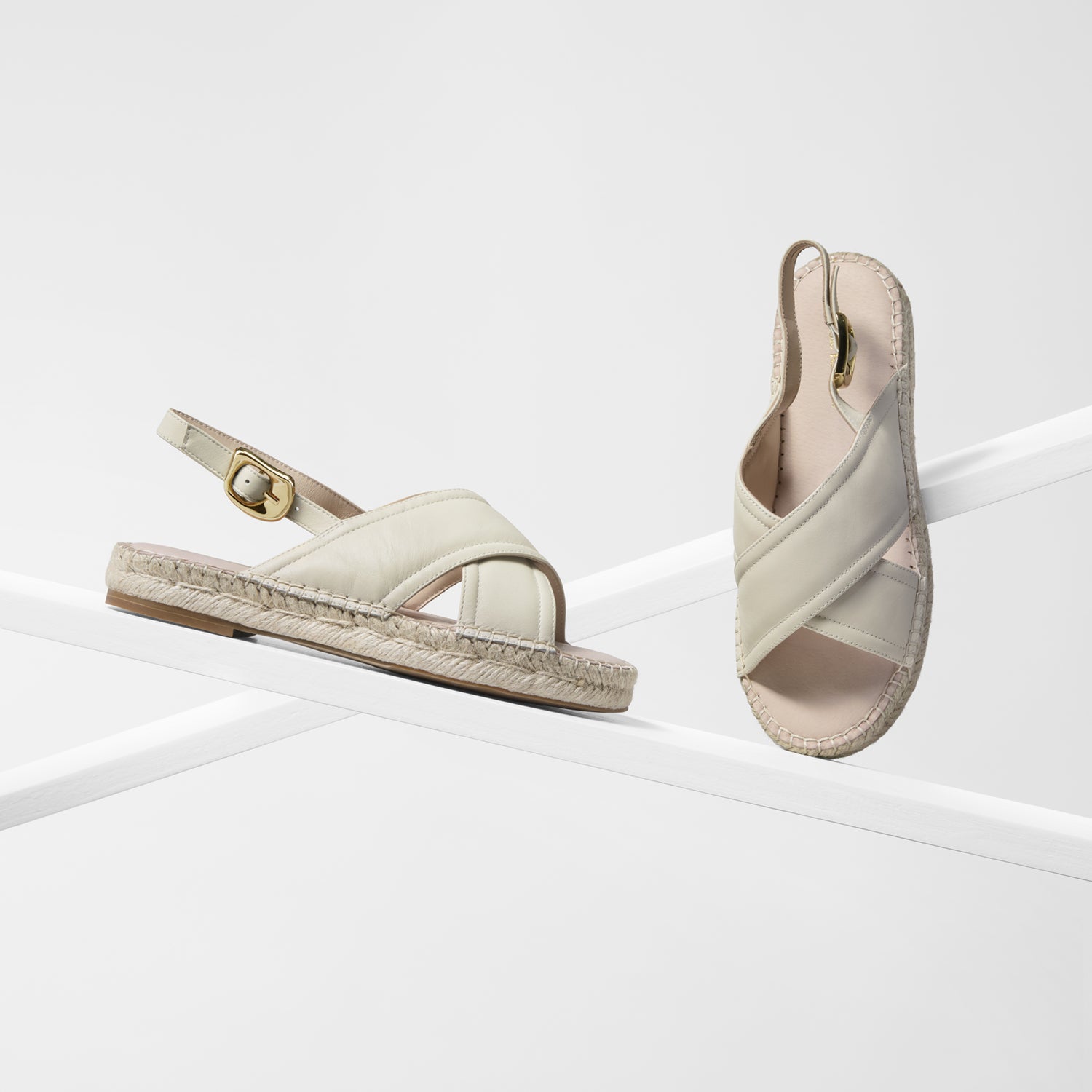 A pair of women's espadrille sandals in a light cream-beige color, featuring wide criss-cross straps, a slingback design, and a braided jute sole. One sandal is shown from the side profile and the other from the top-down view, both resting on a minimalist white geometric platform.