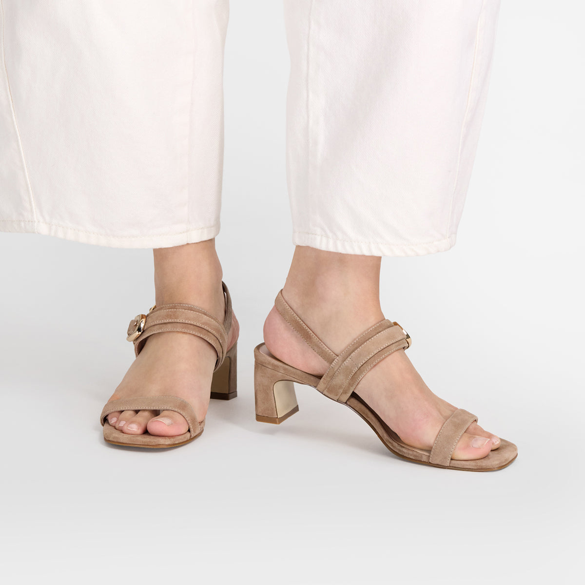 A pair of beige suede block-heel sandals with double front straps and buckle details, shown on a model wearing white pants against a light gray studio background. Made by Nicole Rebstock.