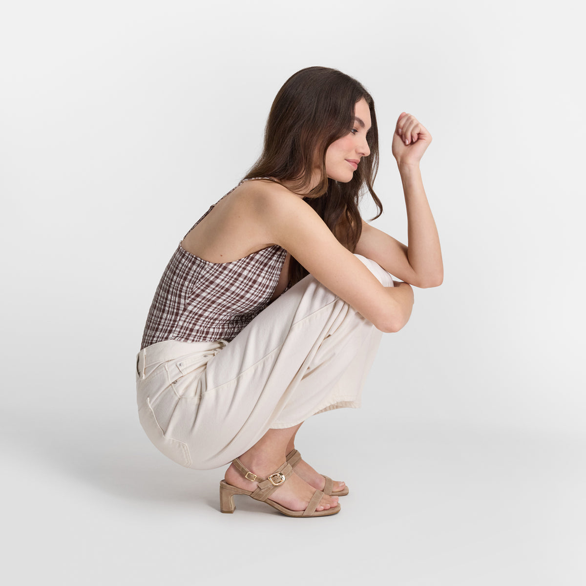 A woman crouches in a studio setting wearing a brown and beige plaid camisole top, cream wide-leg pants, and beige block-heel sandals.