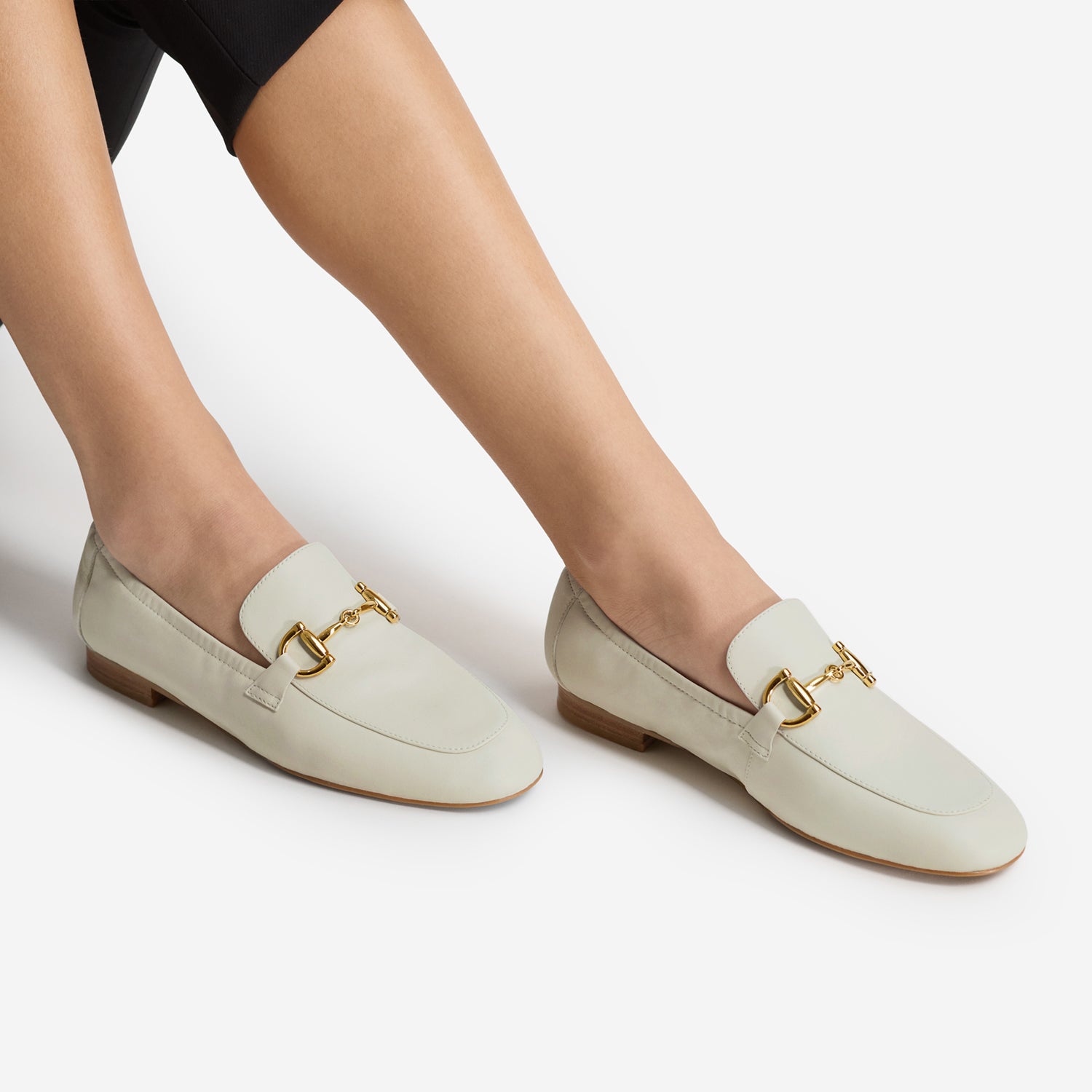 A pair of off-white leather loafers with gold horsebit hardware, shown on a model's feet against a clean white background.