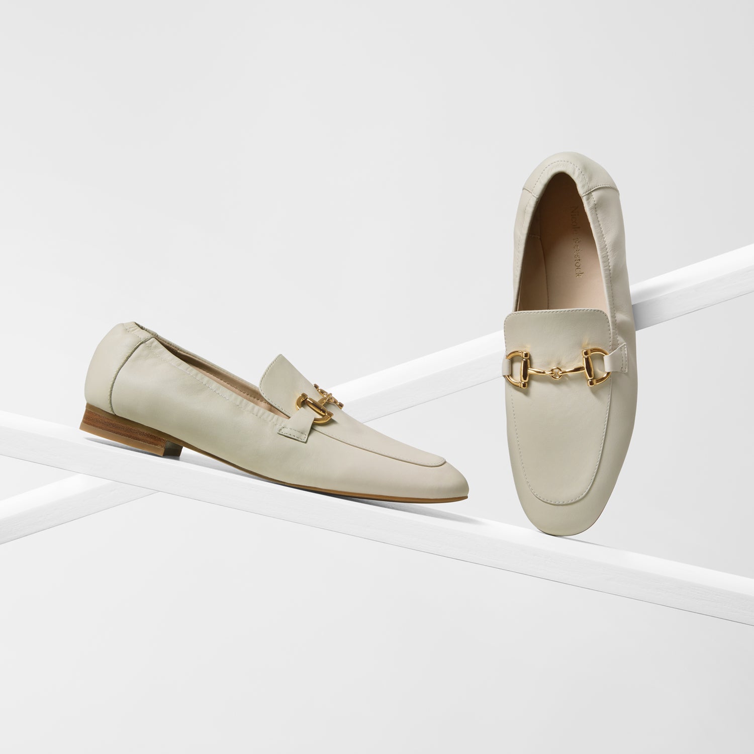 A pair of light beige leather loafers with gold-tone horsebit detailing, displayed on a minimalist white geometric platform against a clean white background.