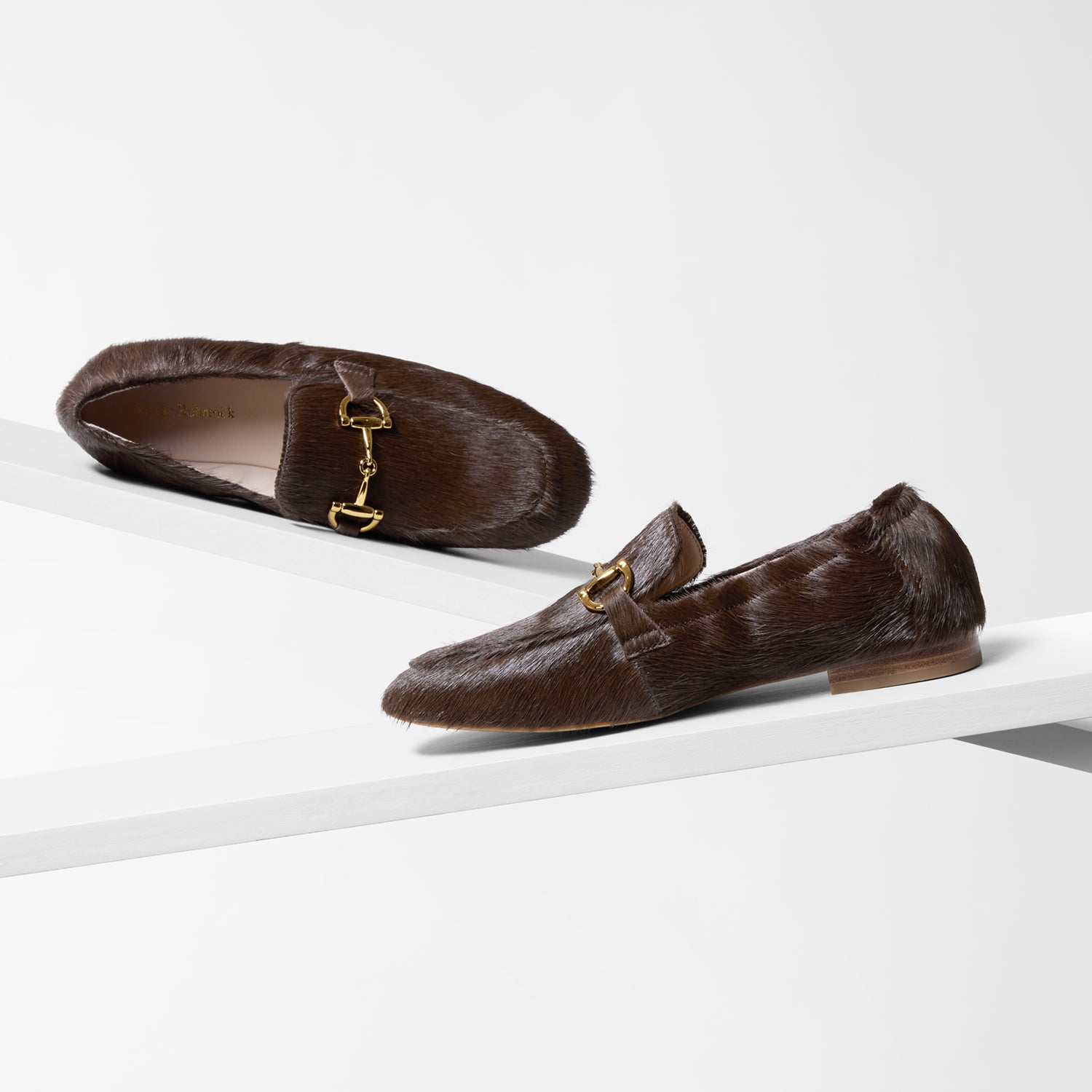 Pair of brown coffee calf hair loafers with gold buckles on a white background