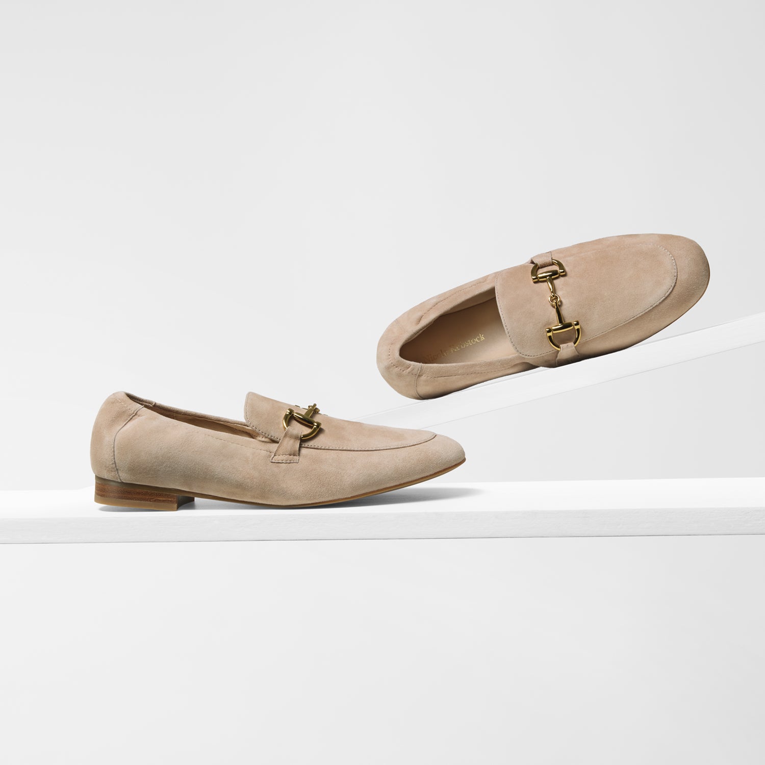 A pair of nude suede penny loafers ny Nicole Rebstock with gold-tone horsebit hardware and elasticated top line, displayed on a clean white platform against a white background.