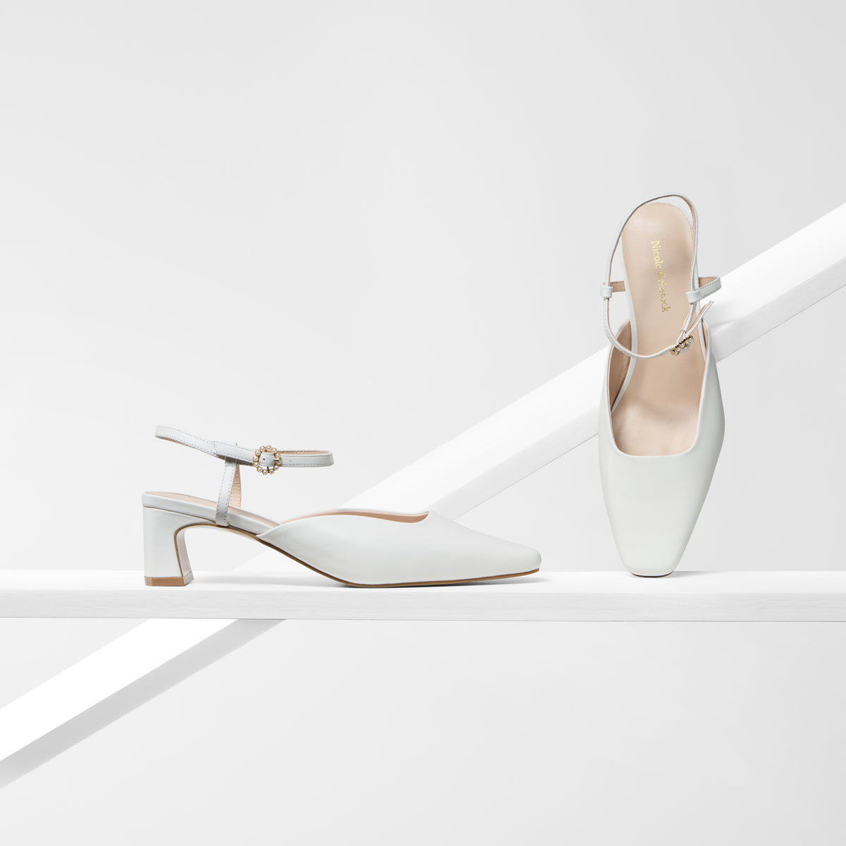 A pair of white pointed-toe heeled mules with ankle straps, one standing upright and the other resting on its side, displayed on a clean white geometric set.