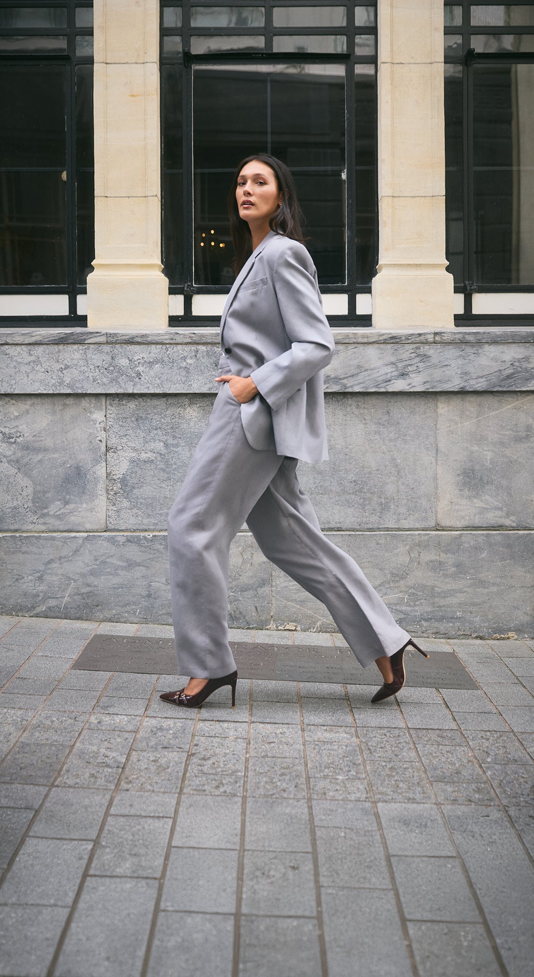 Nash pinot noir patent pump on model walking down the street wearing grey suit.