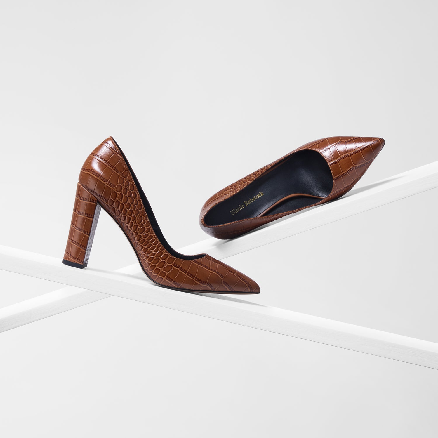 A pair of brown pointed-toe high heels with an embossed crocodile pattern, displayed on a minimalist white geometric platform against a light gray background.