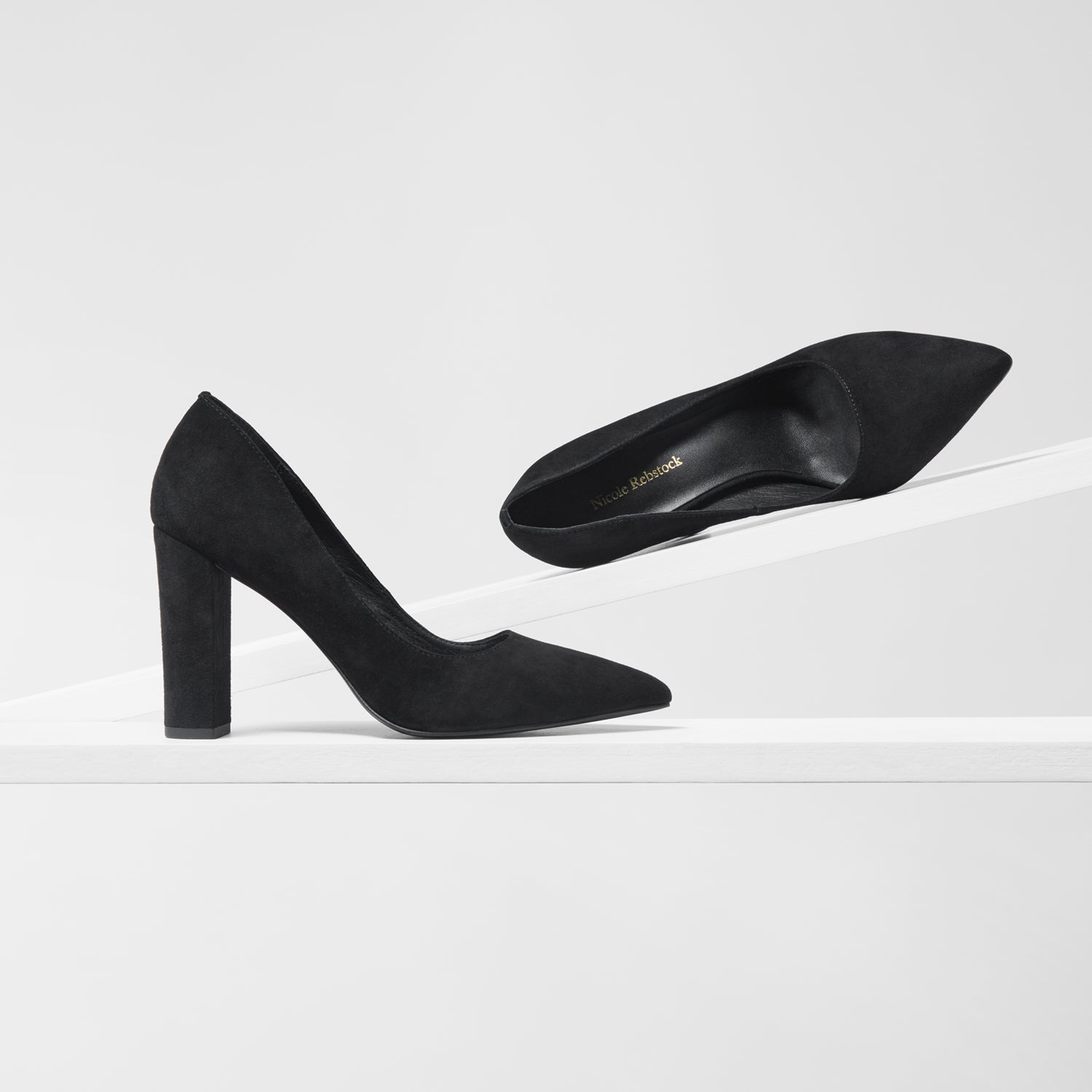 A pair of black pointed-toe high heels made of suede, displayed on a clean white geometric riser against a light gray background.
