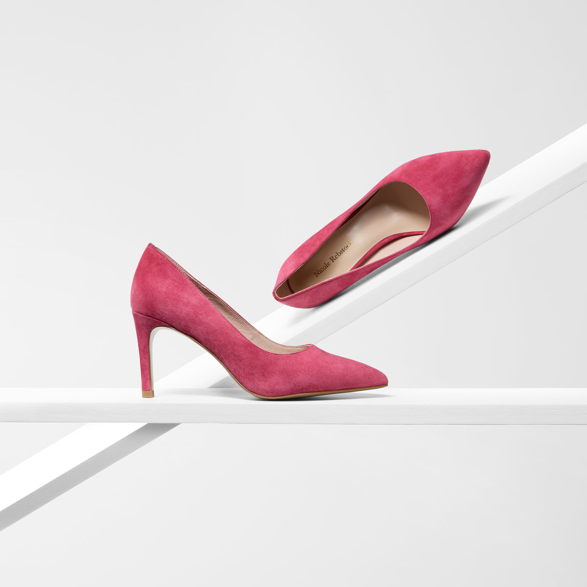 A pair of bright pink suede high-heeled pumps displayed on a minimalist white geometric platform against a light gray background.