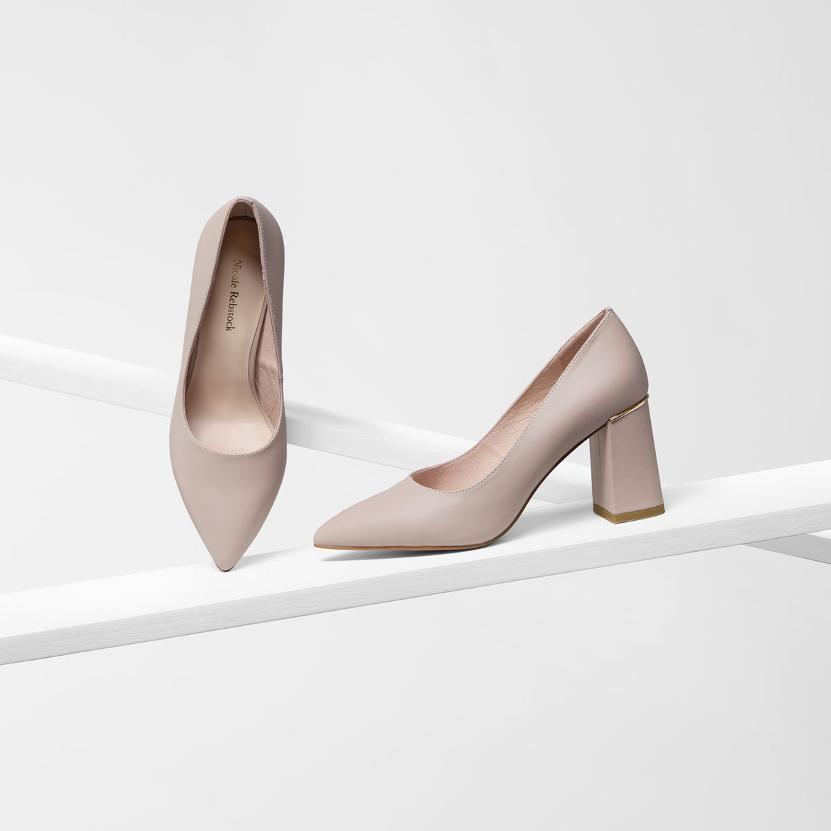 A pair of pointed-toe high heels in a light taupe-beige color, displayed on a minimalist white geometric platform against a clean white background.