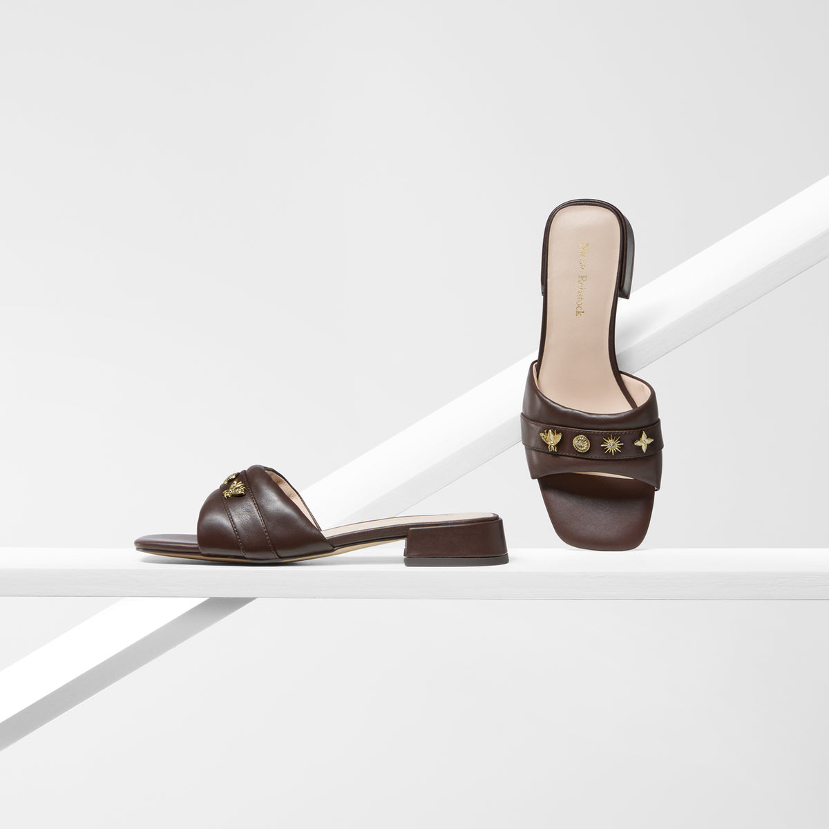 A pair of low heel dark brown leather slide sandals with gold-tone star and crescent moon hardware by Nicole Rebstock, displayed on a minimalist white geometric platform.