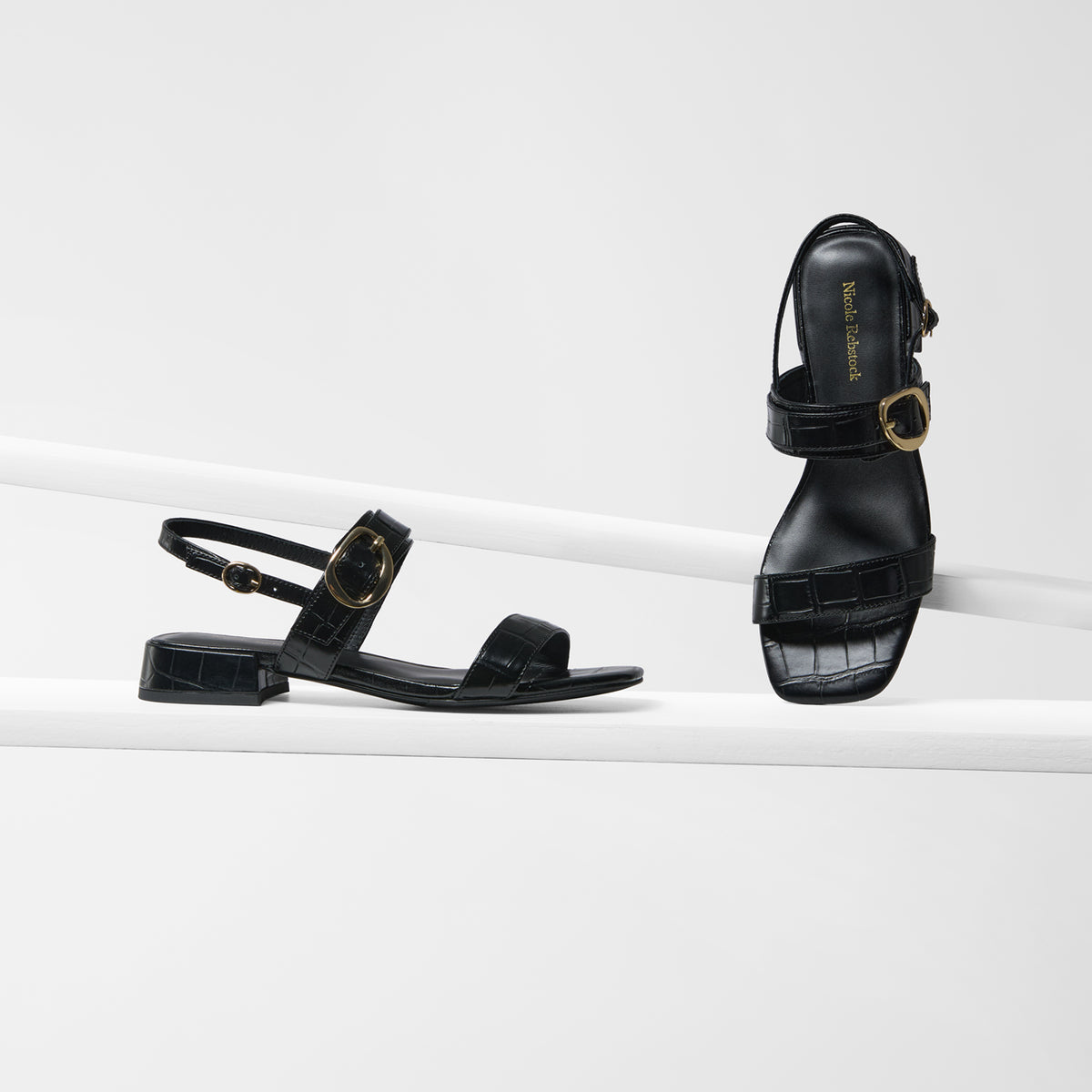 A pair of black leather flat sandals with a low block heel, a single strap with a gold buckle, and a crocodile-embossed toe strap, displayed on a clean white geometric platform.