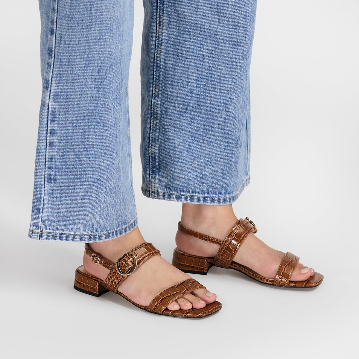 A person wearing brown strappy flat sandals with a crocodile-embossed texture and low block heels. They sandals have square toes and a 2.5cm heel.