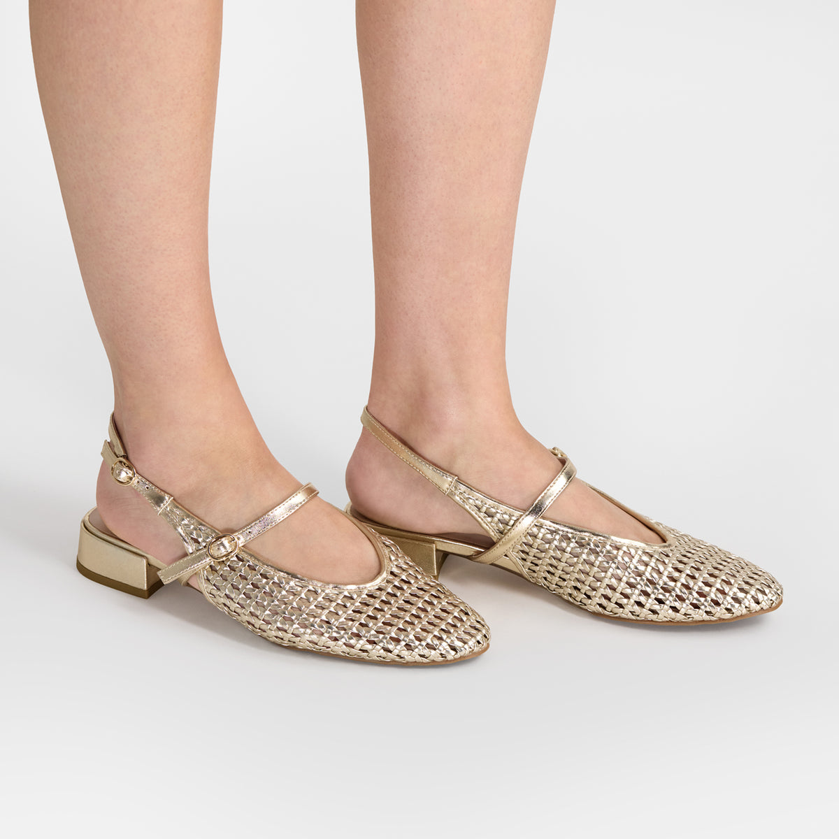 A pair of women's metallic gold flat shoes with a woven mesh upper, ankle straps, and low block heels by Nicole Rebstock, shown on a model's feet against a light gray studio background.