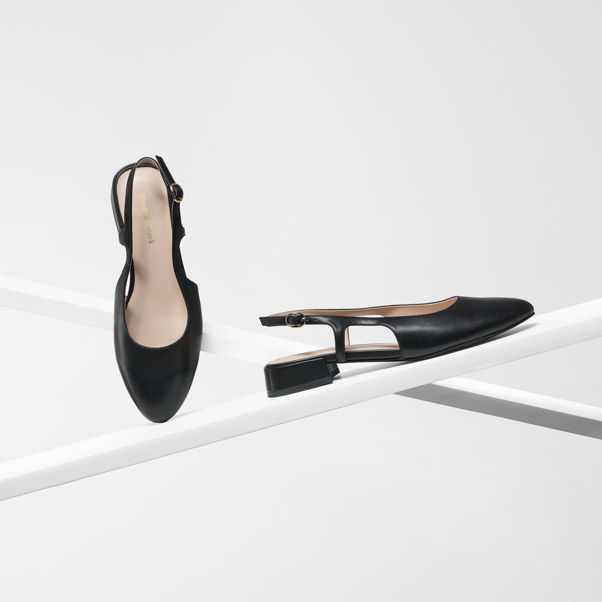 A pair of black leather slingback pumps with block heels, and an almond toe displayed on a minimalist white geometric platform against a clean white background.