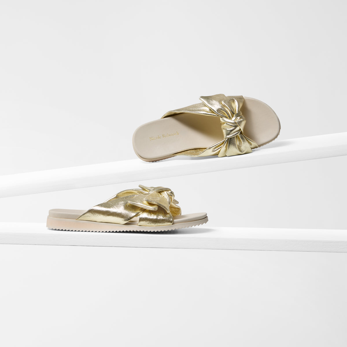 A pair of metallic gold slide sandals with a knotted bow detail on the strap and ivory sole, displayed on a clean white geometric riser.