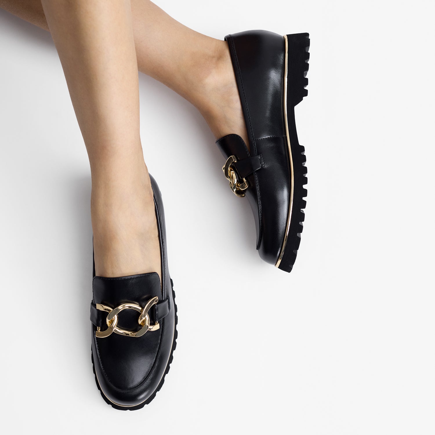 Black loafers with gold chain details worn by a person on a light gray background