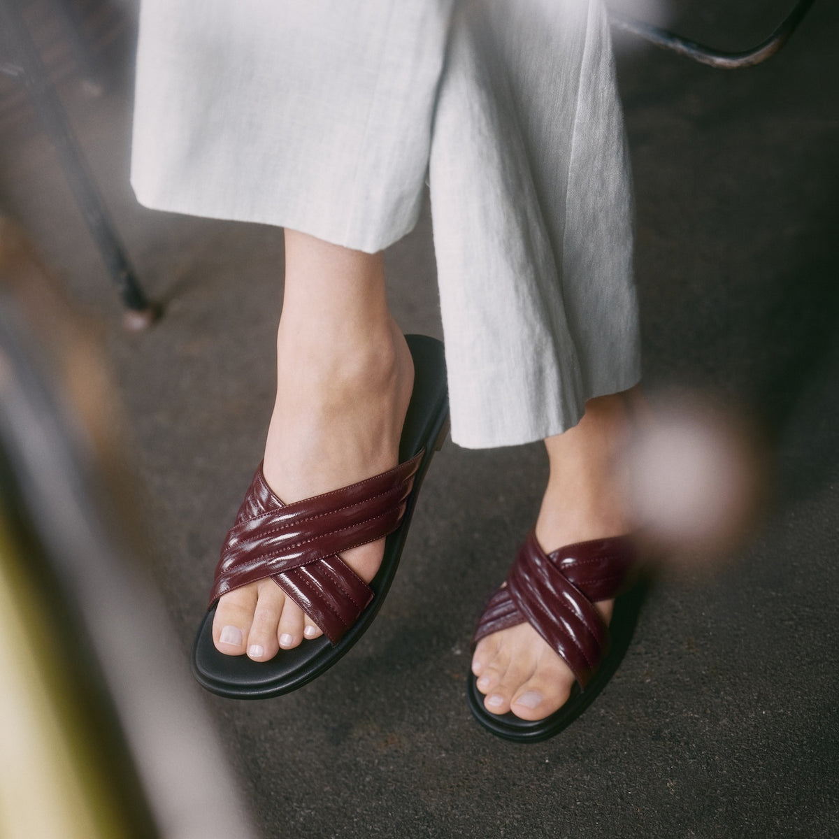 A person wearing flat burgundy crisscross leather slide sandals with black soles by Nicole Rebstock, paired with light gray wide-leg trousers, standing on a dark textured floor.
