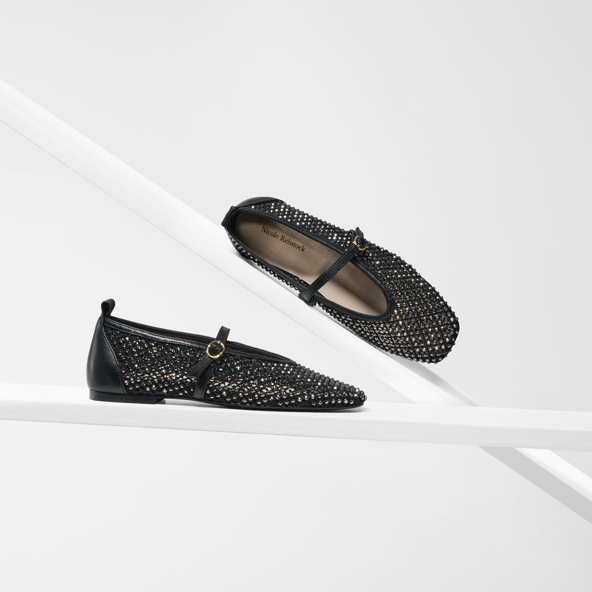A pair of black Mary Jane flats embellished with silver rhinestones, displayed on a clean white geometric background. One shoe is shown from a side profile on a white platform, while the other is positioned diagonally above it to show the interior.