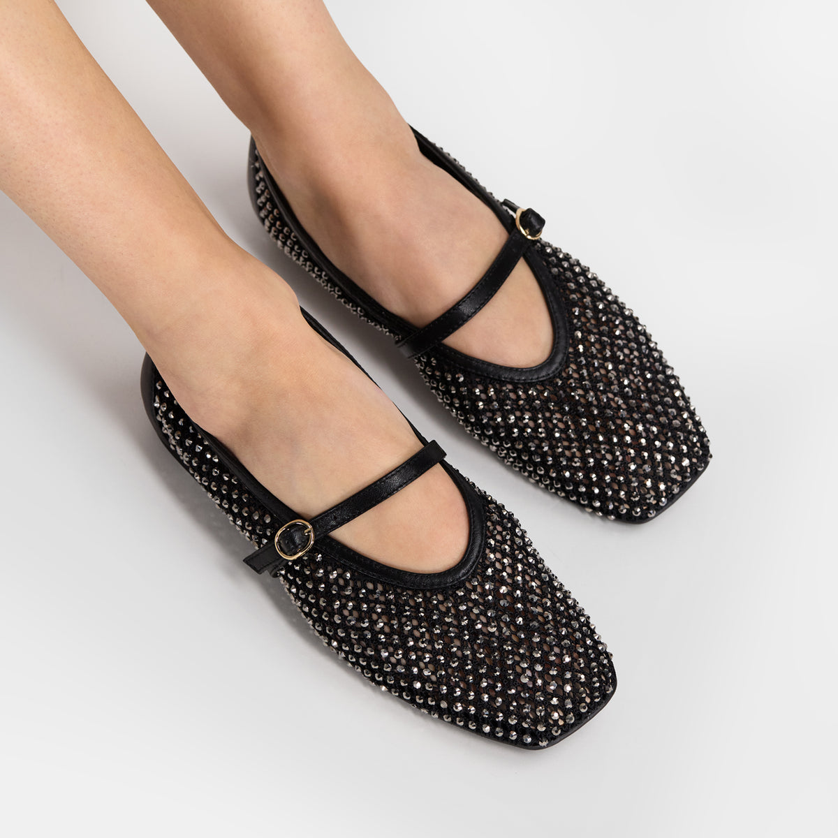 A pair of black Mary Jane flats covered in silver rhinestones, shown on a model's feet against a light gray studio background.