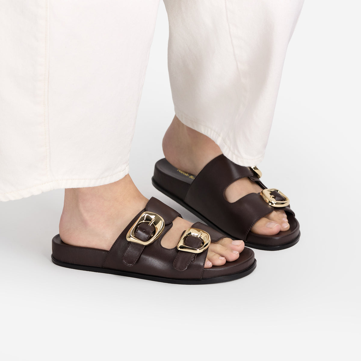 A person wearing dark brown leather slide sandals with double gold-tone buckle straps, paired with white pants, shown against a clean white studio background.