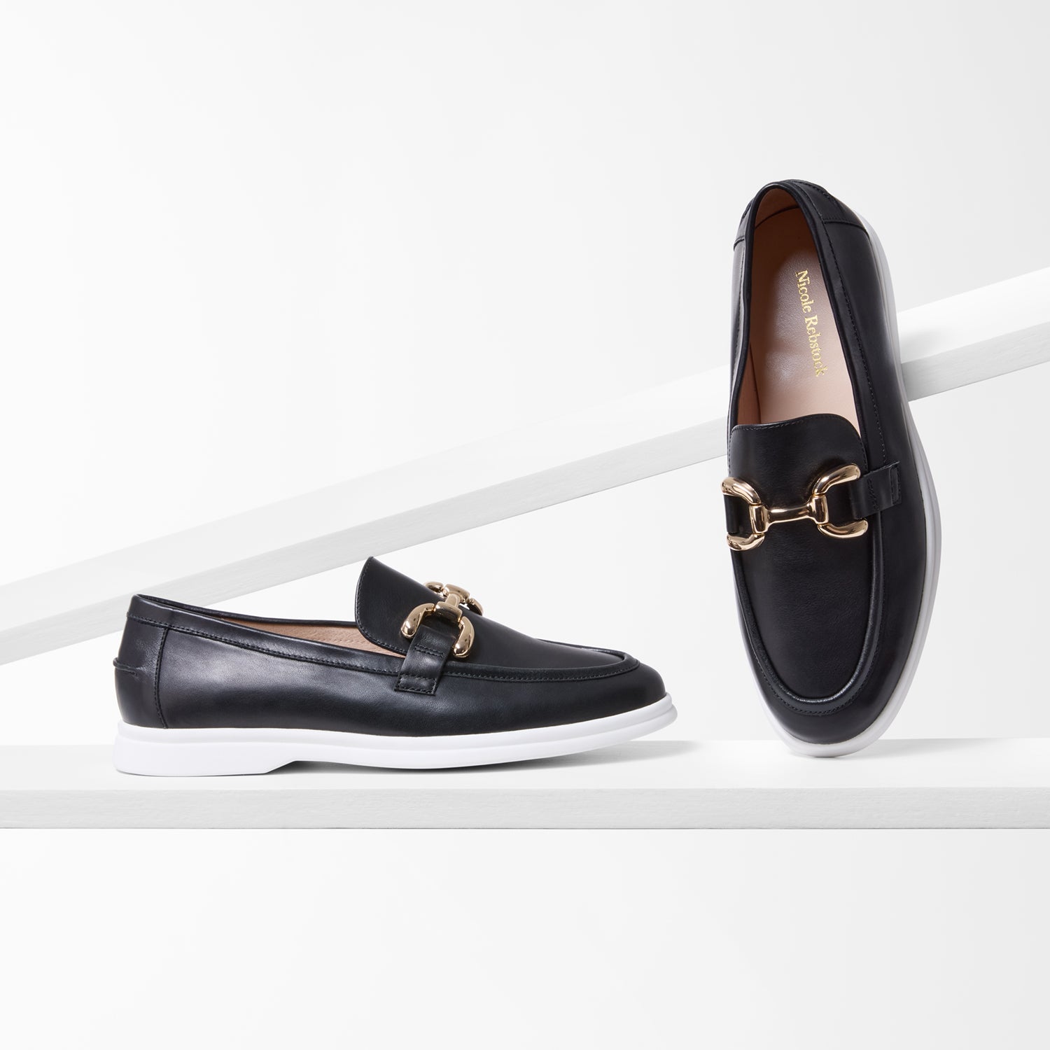 Black loafers with gold buckles on a white background by Nicole Rebstock