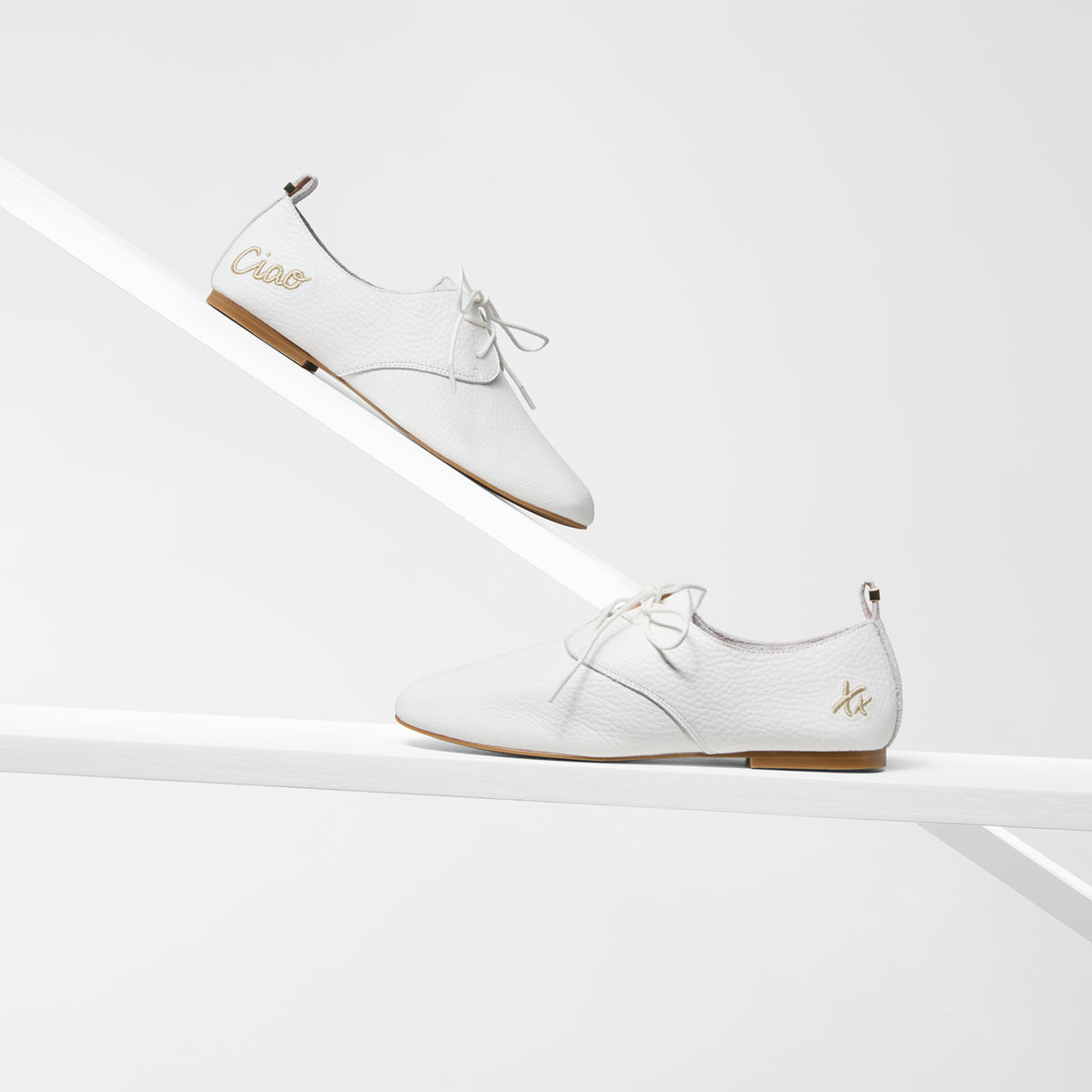A pair of white pebbled leather shoes with laces, displayed on a minimalist white geometric shelf. One shoe is positioned on a diagonal riser above, and the other is on a lower shelf.