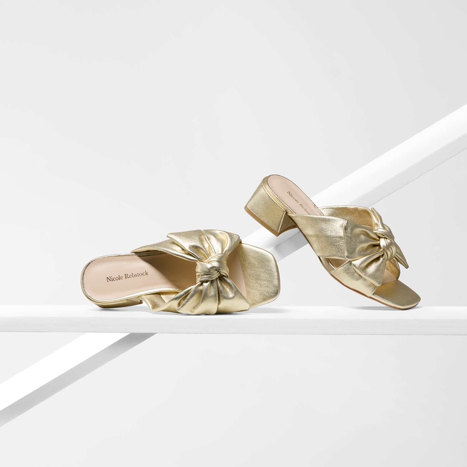 A pair of metallic gold slide sandals with a knotted bow detail on the strap, displayed on a minimalist white geometric platform.